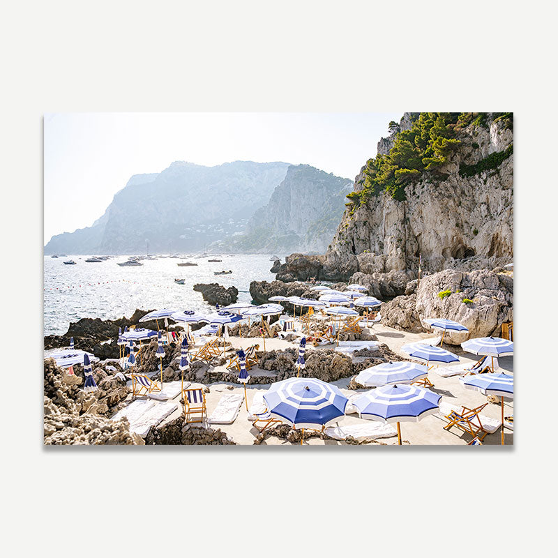 Beach scene with blue umbrellas and chairs on a rocky shore with mountains in the background.