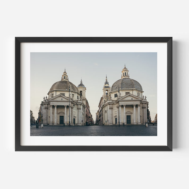 A framed art print featuring a symmetrical photograph of two classical domed churches under a pale sky, presented in a black frame with a white mat against a light gray background.