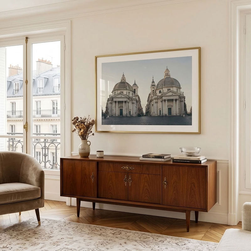 A framed art print depicting classical domed cathedral architecture, displayed on a cream wall above a mid-century modern wooden sideboard in a beautifully styled living room.