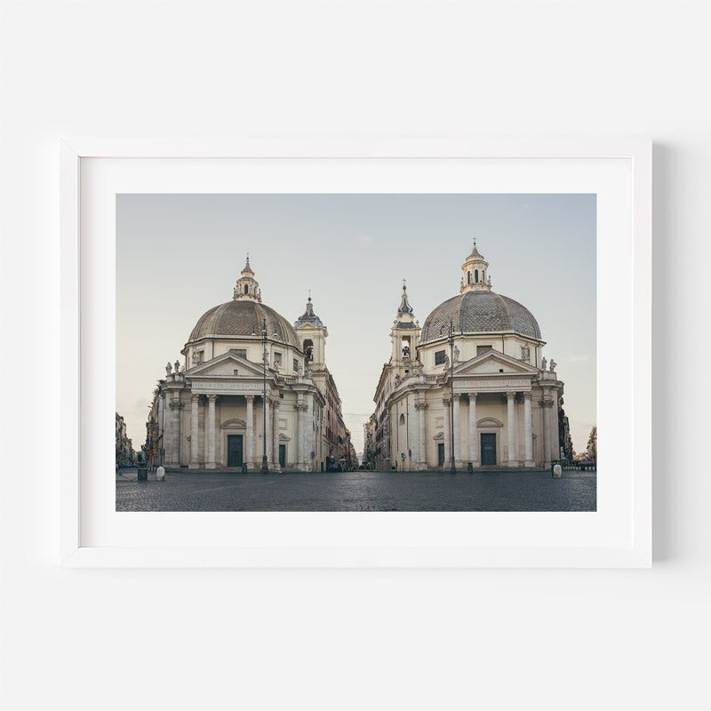 A framed photographic print of two symmetrical baroque-style domed churches in an open square, shown against a clean white background.
