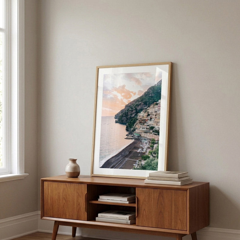 A framed vertical art print depicting a coastal cliffside village scene with warm sunset tones, displayed on a mid-century modern wooden console table in a neutral-toned room.