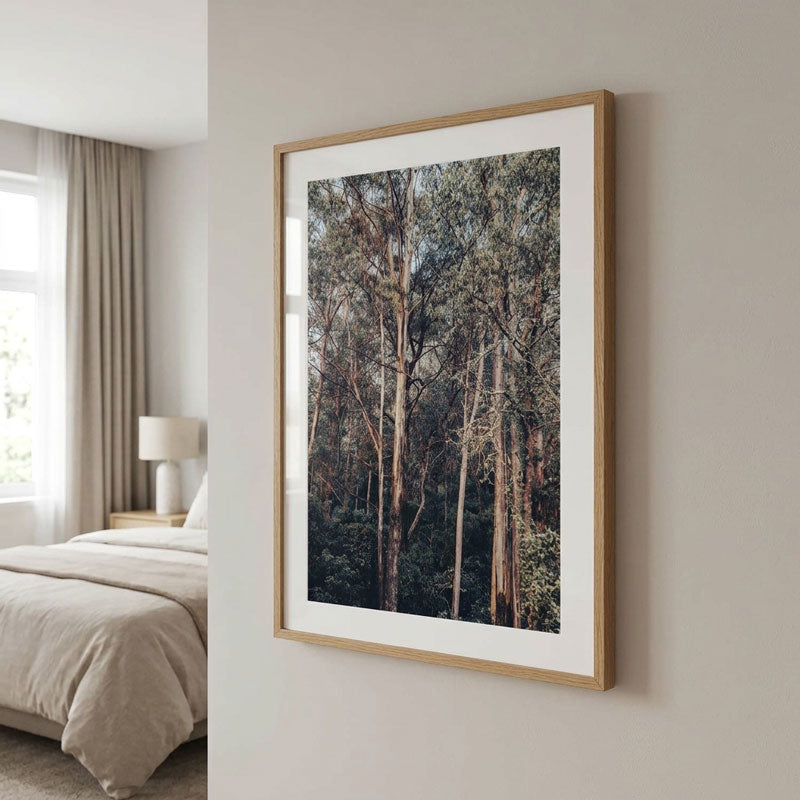 A framed vertical art print depicting a dense forest of tall trees, styled on a light beige wall in a modern bedroom setting.
