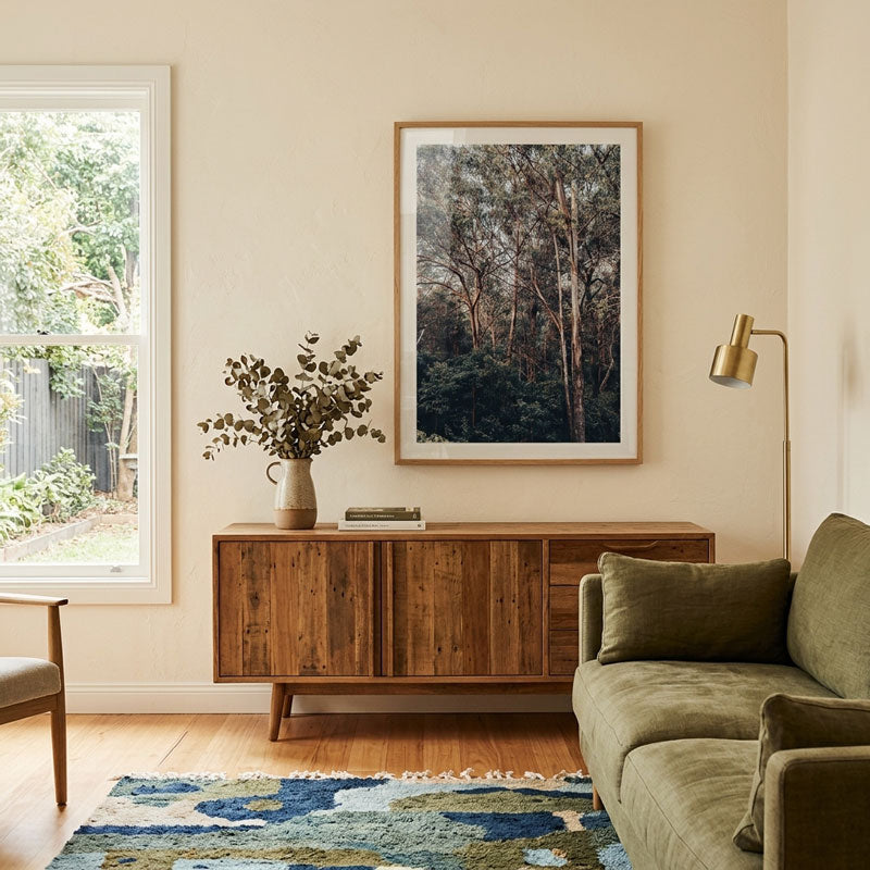 A framed forest art print hangs on a beige wall above a wooden sideboard in a styled living room with an olive green sofa, a window, and a blue patterned rug.