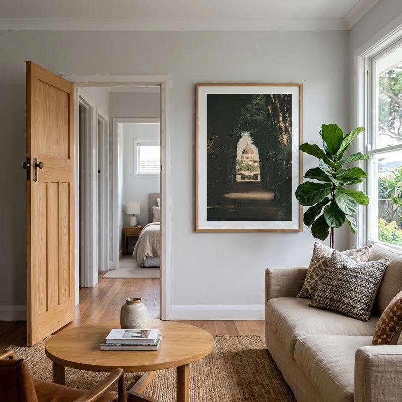 A framed art print depicting a dark forest path with a bright window of light, styled in a modern living room next to a beige sofa, a wooden coffee table, and a green plant.