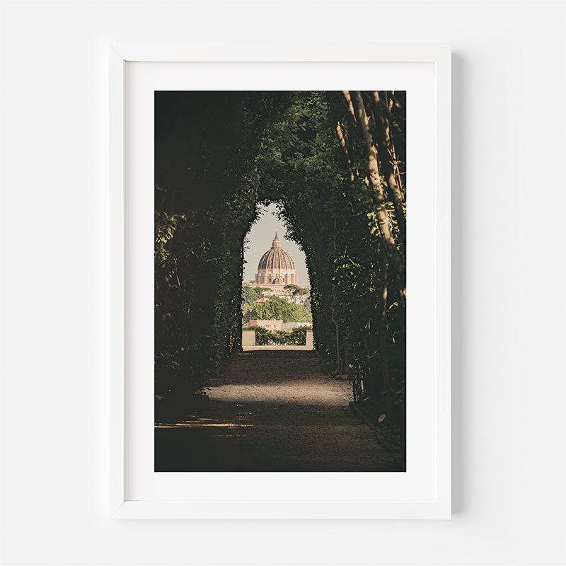A framed art print featuring a scenic photograph of a domed cathedral visible through a tree-lined path, presented in a white frame against a light gray wall.