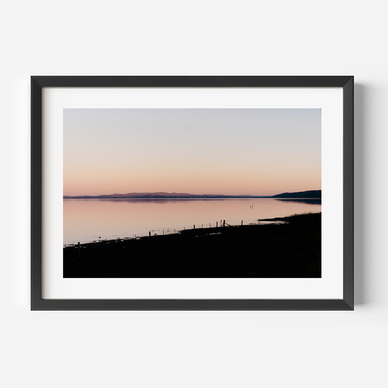 Framed photograph of a sunset over a body of water with a silhouette of a fence and landscape.