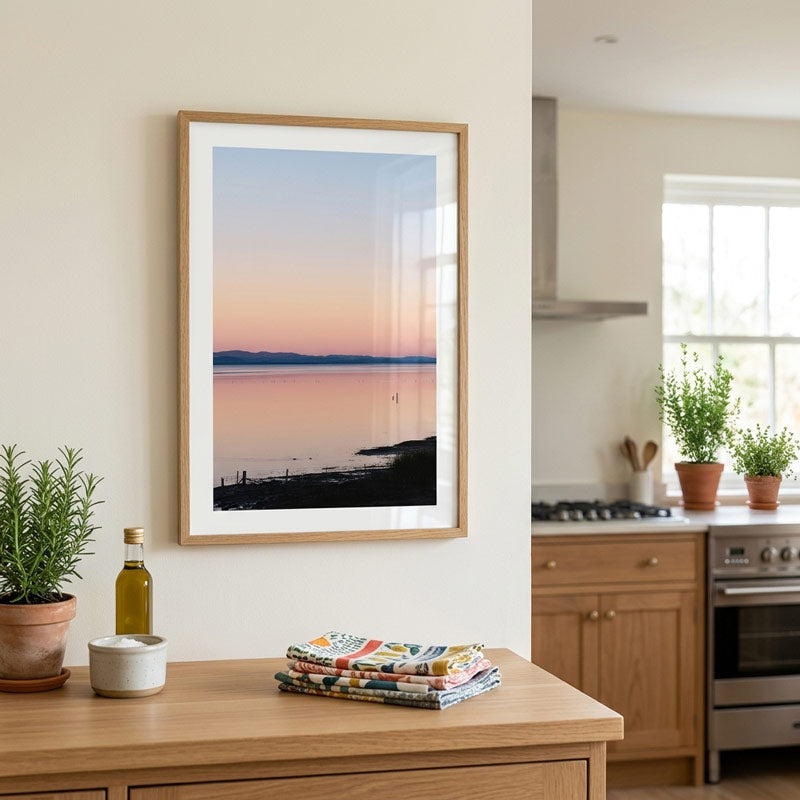 A framed vertical landscape art print of a sunset over water, styled on a cream wall above a wooden console table in a modern kitchen setting.