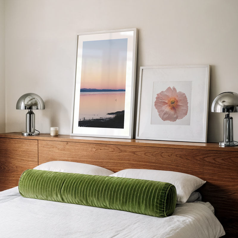 Green cylindrical pillow on a bed with framed artwork and lamps in the background.