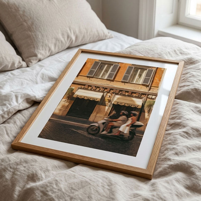 A framed art print depicting a vintage scooter scene in front of an orange building, styled on a bed with neutral-colored linen bedding.