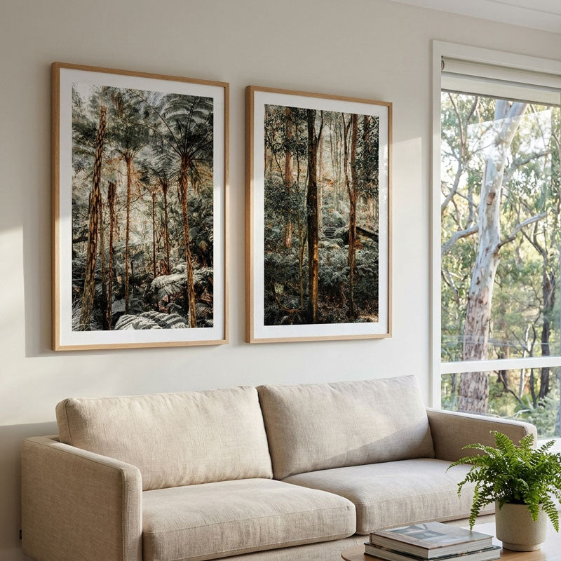Two vertical framed forest art prints hang on a light beige wall above a matching beige sofa, with a window showing trees outside and a potted plant on a table in the foreground.