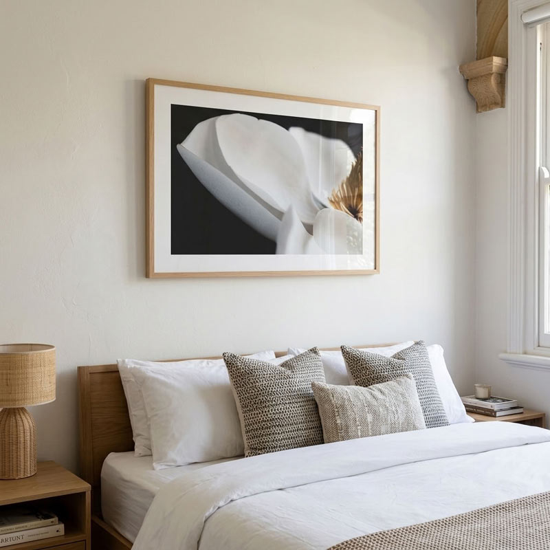 A framed black and white floral art print hangs on a beige wall above a neatly made bed with white linens and textured gray pillows in a bright, modern bedroom.