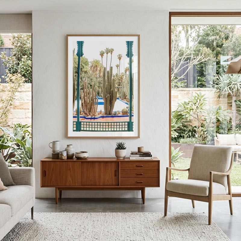 A framed art print depicting a desert cactus garden with teal architectural elements, displayed on a white wall above a mid-century modern wooden sideboard in a bright, contemporary living room.