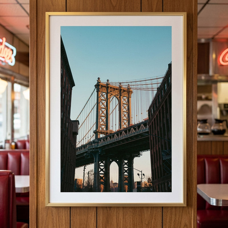 A framed art print of the Manhattan Bridge in New York City, featuring a white mat and a gold frame, displayed on a wooden wall in a restaurant setting.