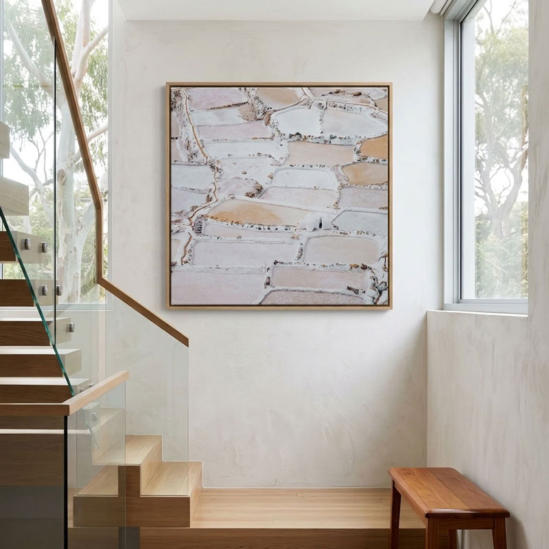 A large framed abstract landscape painting with geometric fields in beige, white, and brown tones, displayed on a white wall in a modern staircase hallway.
