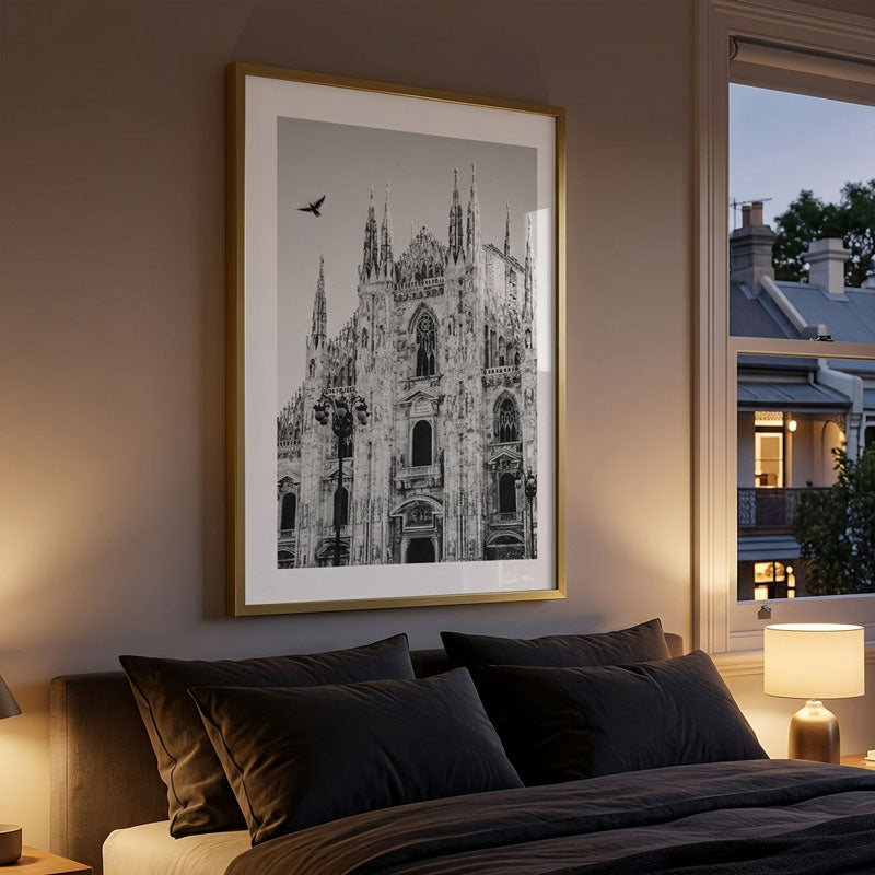 A framed black and white architectural art print of the Milan Cathedral hangs on a warm beige wall above a bed with dark pillows in a styled bedroom setting.