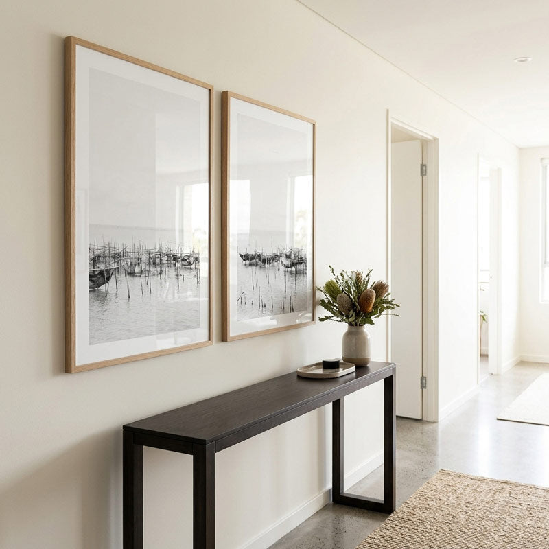 Two large black and white framed art prints with light wood frames hang on a cream wall above a dark console table in a bright, modern hallway.