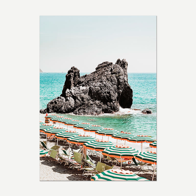 Beach scene with rows of striped umbrellas and lounge chairs near a rocky outcrop in the ocean.