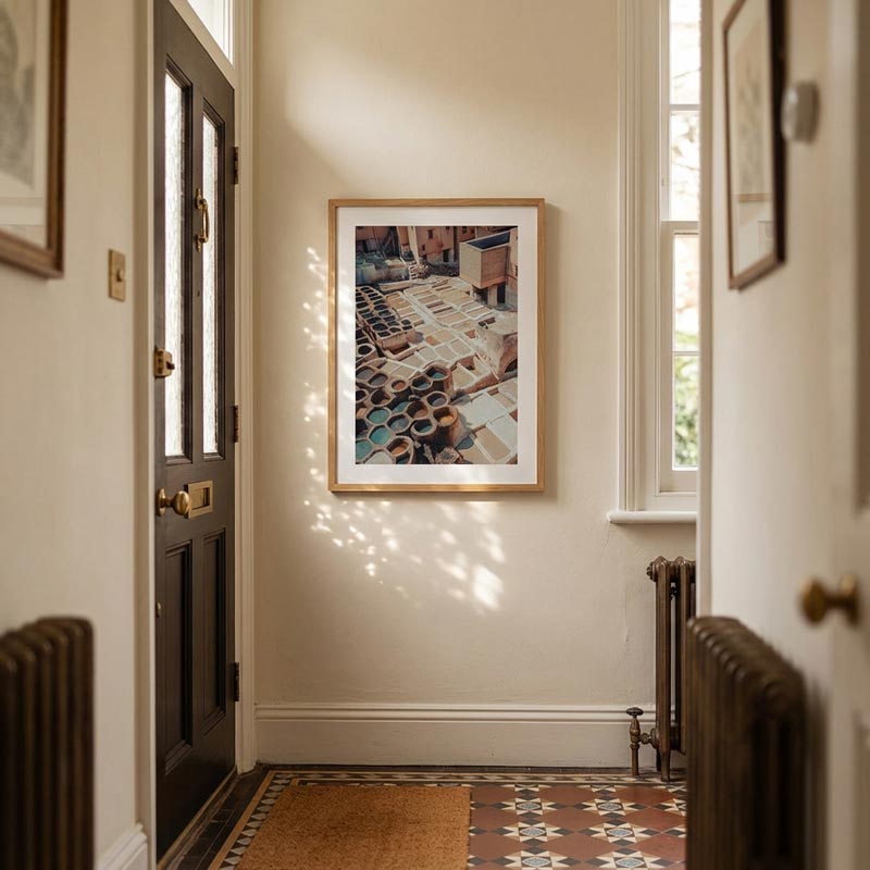 A framed art print depicting a colorful pottery workshop hangs on a warm beige wall in a cozy, sunlit hallway with a patterned tile floor and dark wood door.