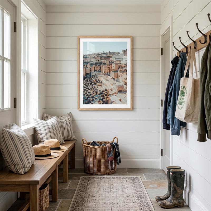 A framed art print depicting a rustic courtyard with stone paving and terracotta tiles, hanging on a white shiplap wall in a bright, modern entryway with a wooden bench, striped pillows, a wicker basket, and hanging jackets.