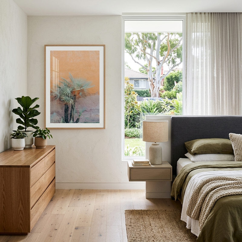 A framed abstract landscape art print featuring a desert scene with a cactus and palm tree, styled in a modern, bright bedroom next to a wooden dresser and window.