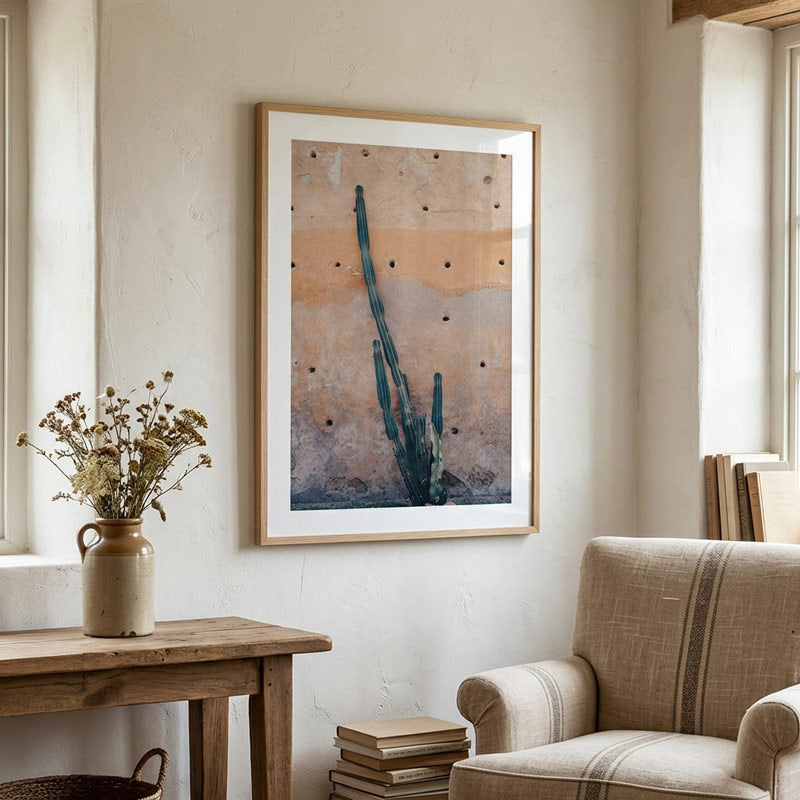 A framed abstract art print featuring a stylized cactus on a textured beige and orange background, hanging on a cream-colored wall in a styled room with a wooden table, vase of dried flowers, and a beige armchair.
