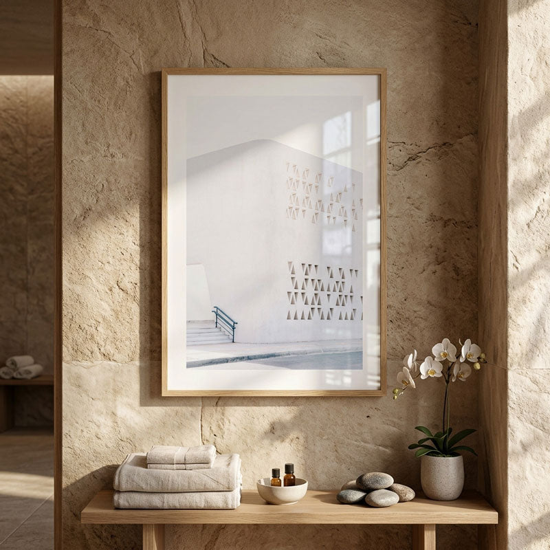 A framed minimalist art print depicting a white modern building with black architectural lines, hanging on a textured beige stone wall above a wooden console table styled with folded towels, a bowl, and a potted orchid.