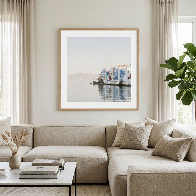 A framed art print depicting a scenic harbor with colorful boats and distant mountains, displayed on a light beige wall above a modern beige sectional sofa in a bright, contemporary living room.