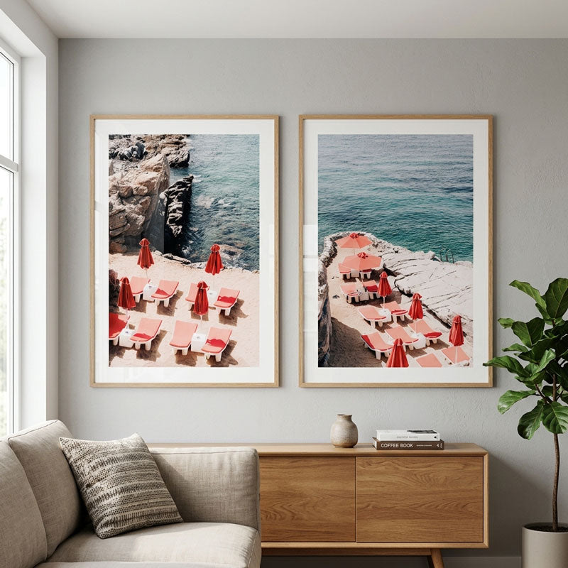 Two framed coastal beach prints with red lounge chairs and umbrellas hang on a light gray wall above a wooden console table in a modern living room setting.