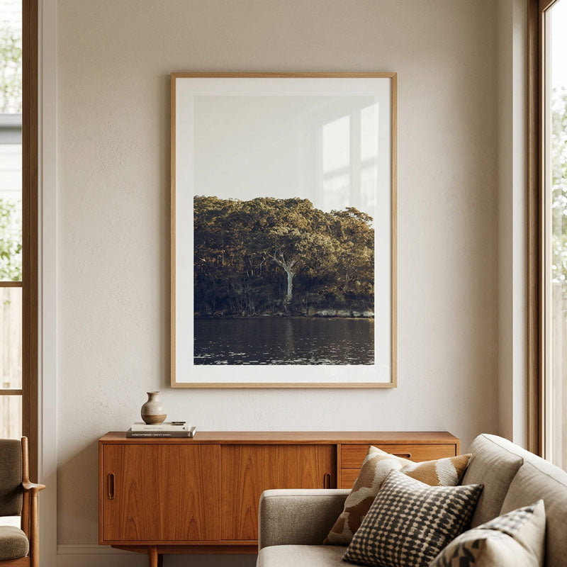 A framed art print depicting a scenic forest by a lake, styled in a modern living room setting above a mid-century wooden sideboard and next to a beige sofa.