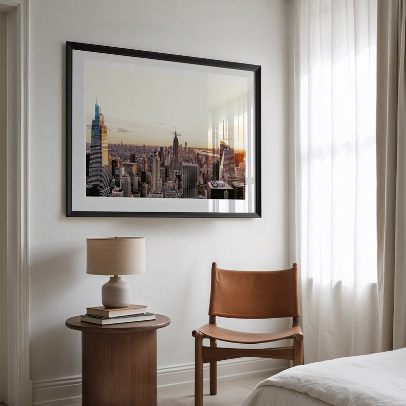 Framed cityscape artwork on a wall above a side table with a lamp and books, next to a brown chair.
