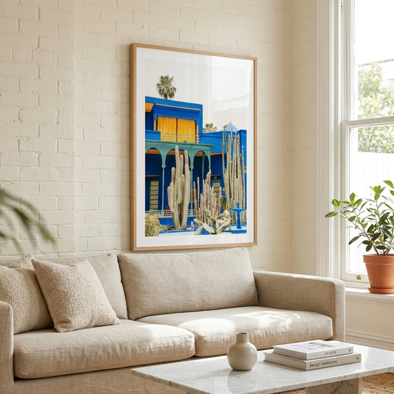 A framed art print featuring a vibrant blue desert building and cacti, displayed on a white brick wall above a beige sofa in a bright, modern living room.