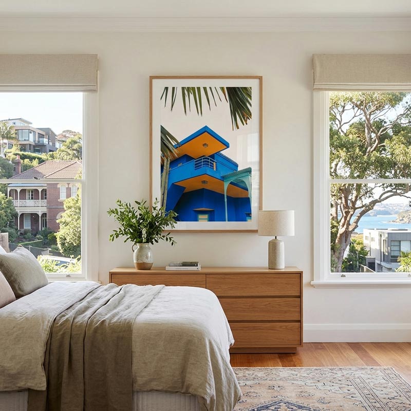 A framed art print featuring a vibrant blue and yellow house with palm leaves, displayed on a beige wall above a wooden dresser in a modern, sunlit bedroom.
