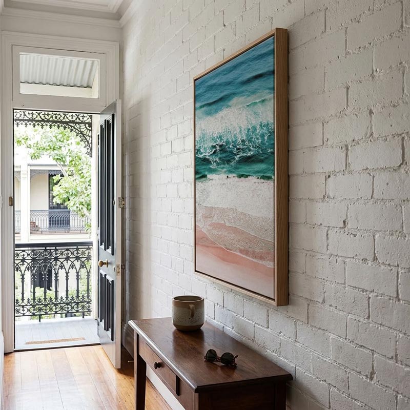 A framed vertical art print depicting a turquoise ocean wave crashing onto a sandy beach, hung on a white brick wall above a dark wood console table in a bright, modern room.