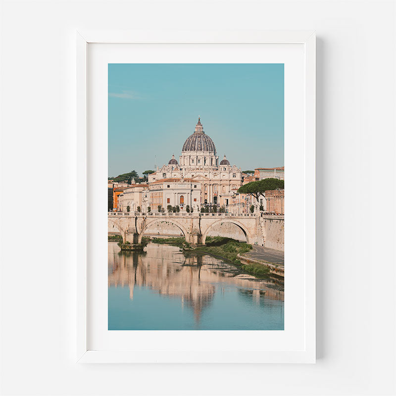 A framed art print depicting St. Peter's Basilica in Rome, with the bridge and river reflection below, presented in a white frame against a light gray wall.