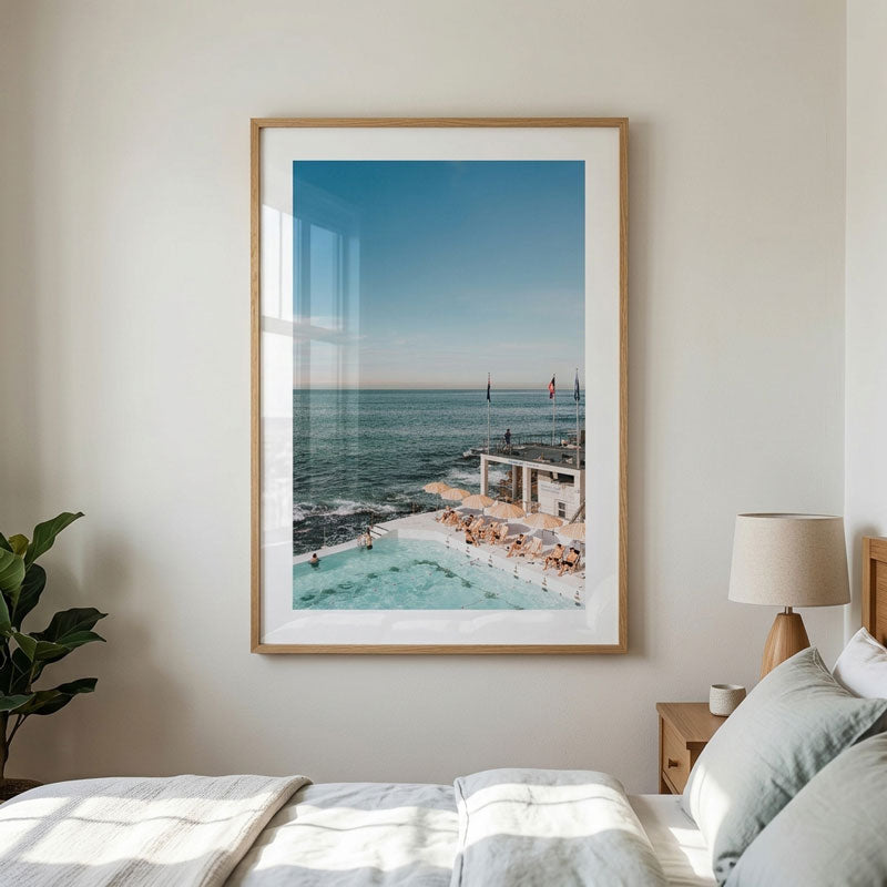 A framed beach and poolside print hanging on a light beige wall above a bed in a modern bedroom. The artwork shows a coastal scene with blue water, a swimming pool, and a sunlit deck.