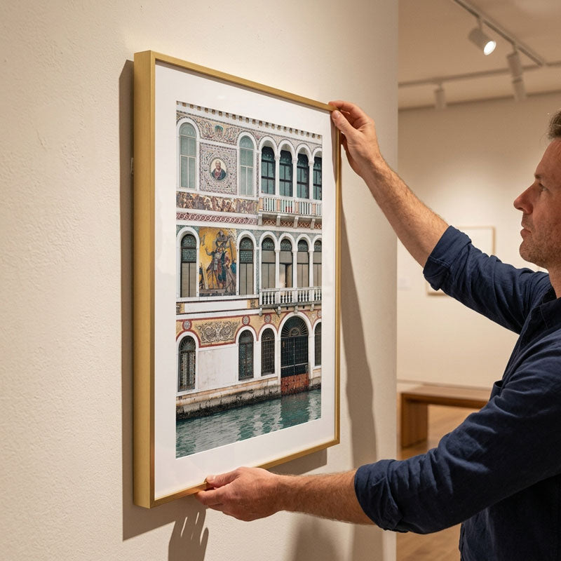 A man in a navy blue shirt is shown holding a framed art print of a Venetian building facade, which features a white mat and a light wood frame, displayed on a gallery wall.