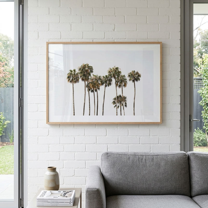 Framed artwork of palm trees on a white brick wall above a gray sofa.