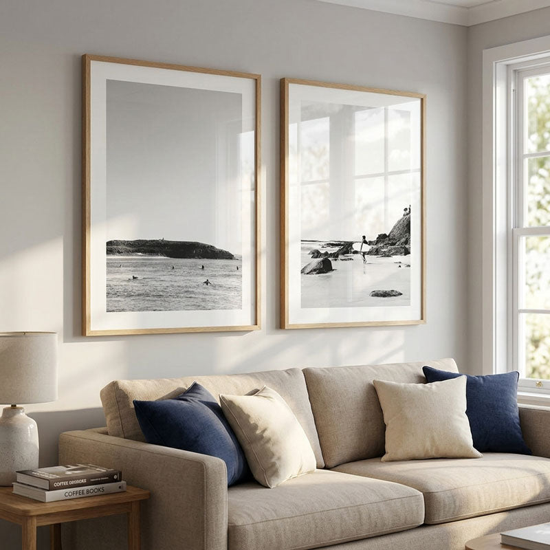 Two vertical framed black-and-white beach landscape prints with light wood frames hang on a light gray wall above a beige sofa with navy and cream pillows in a bright living room.