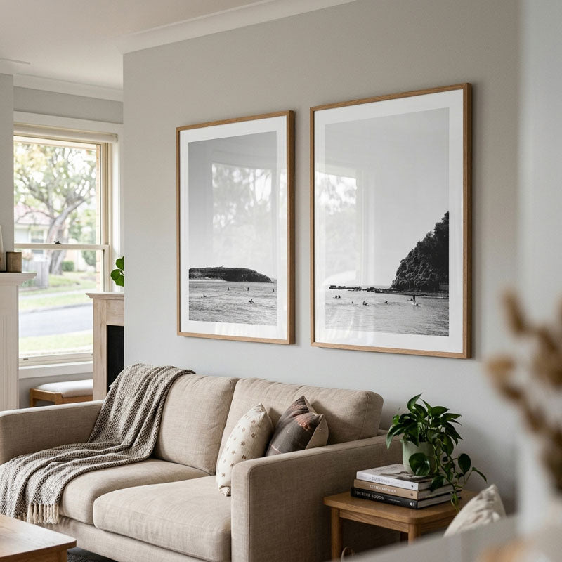 Two framed black and white beach landscape prints with light wood frames hang on a light gray wall above a beige sofa in a styled living room.