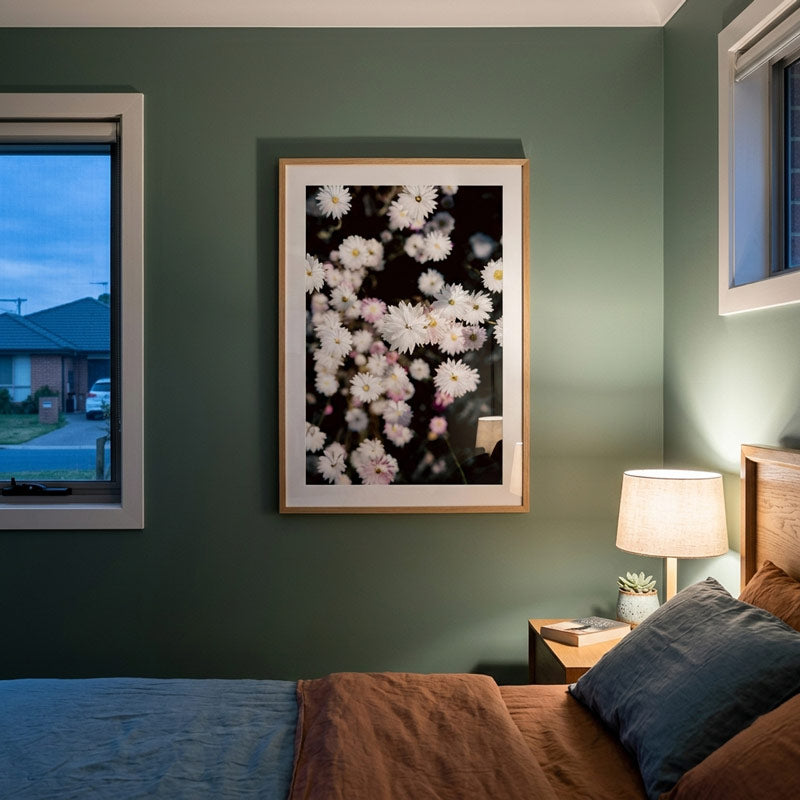 A framed floral art print featuring white and pink daisies on a dark background, hung on a sage green wall above a bed in a styled bedroom.