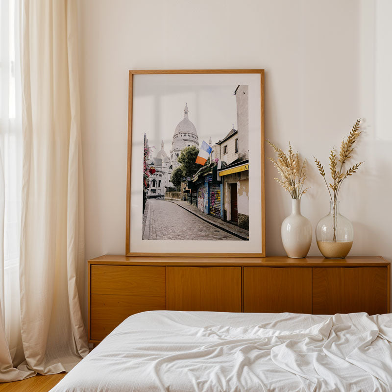 Framed artwork on a wooden dresser with white curtains and a bed in the foreground