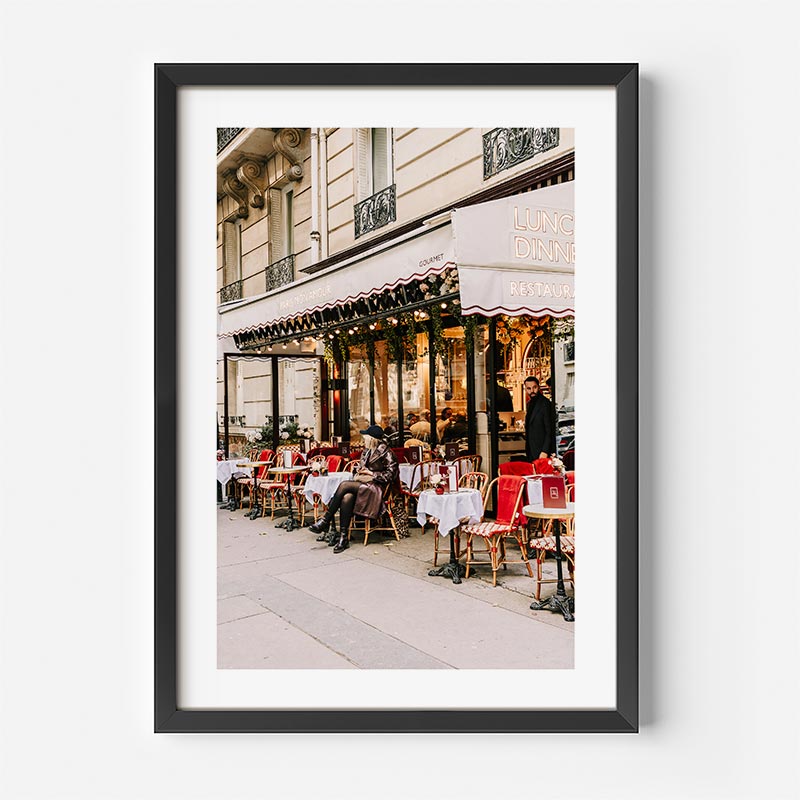 Framed photograph of a street scene with people sitting at outdoor cafe tables in front of a building with a sign.