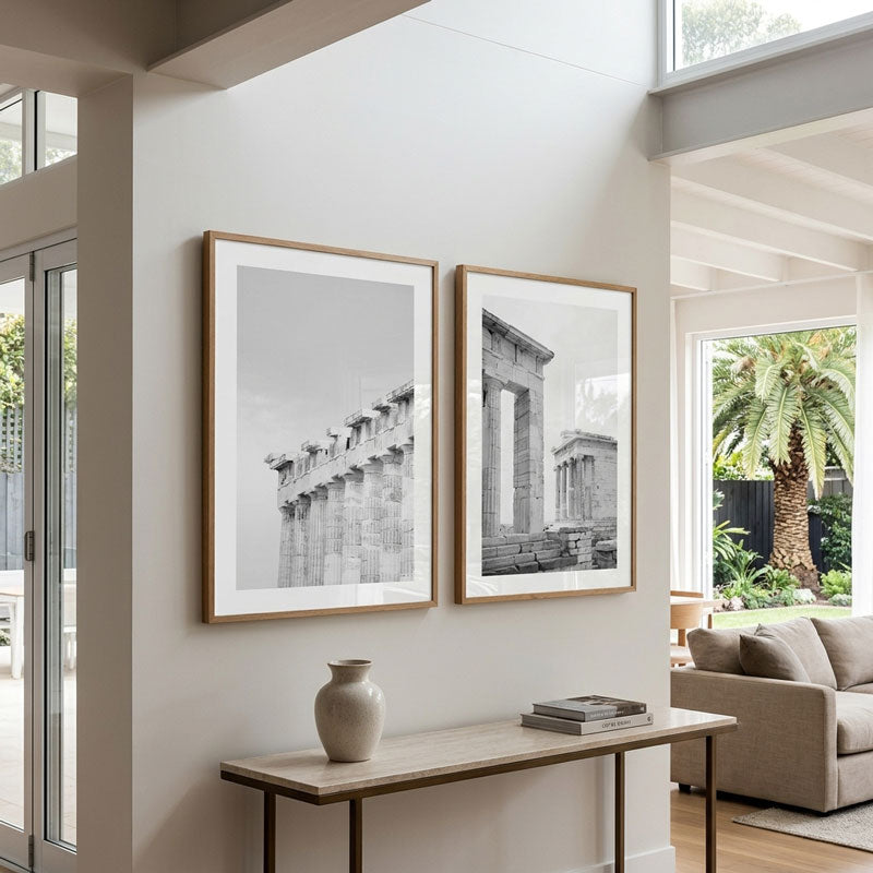 Two framed black and white architectural art prints hang on a white wall in a modern, sunlit living space with a console table and sofa below.