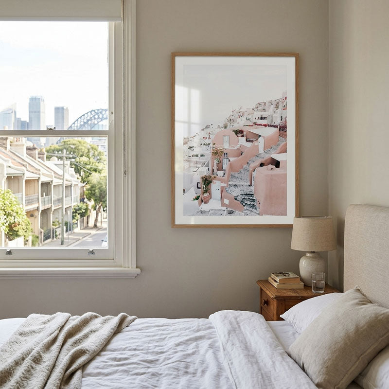 A framed art print featuring a pink and white architectural scene, displayed on a neutral beige wall in a modern bedroom setting next to a bed and a window with a city view.