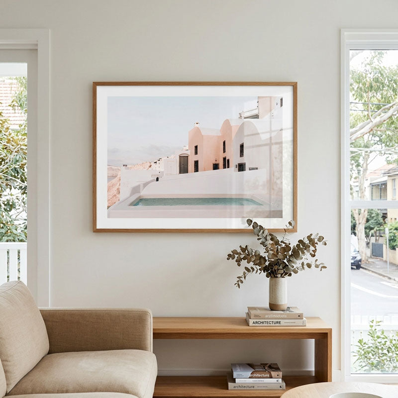 A framed art print depicting a Mediterranean-style white building with a pink terrace and pool, displayed on a light gray wall above a wooden console table in a modern living room setting.