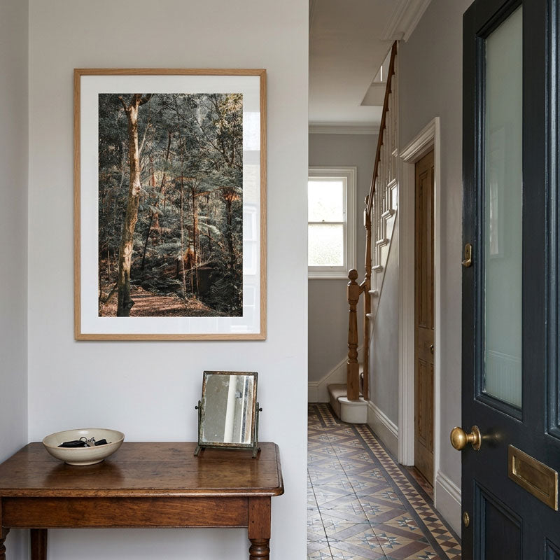 A framed forest landscape print hangs on a white wall in a hallway above a wooden console table. The artwork shows a wooded path with tall trees in muted earthy tones, surrounded by a white mat and light wood frame.
