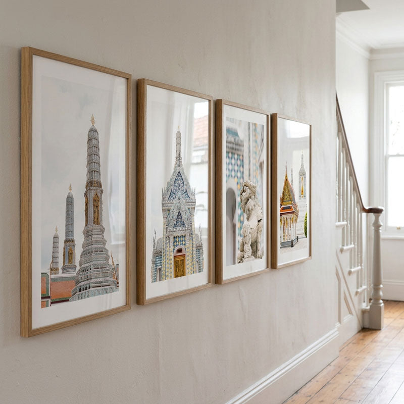 Four framed art prints depicting Thai temple architecture hang in a row on a light beige wall in a bright hallway with wooden floors and a staircase.