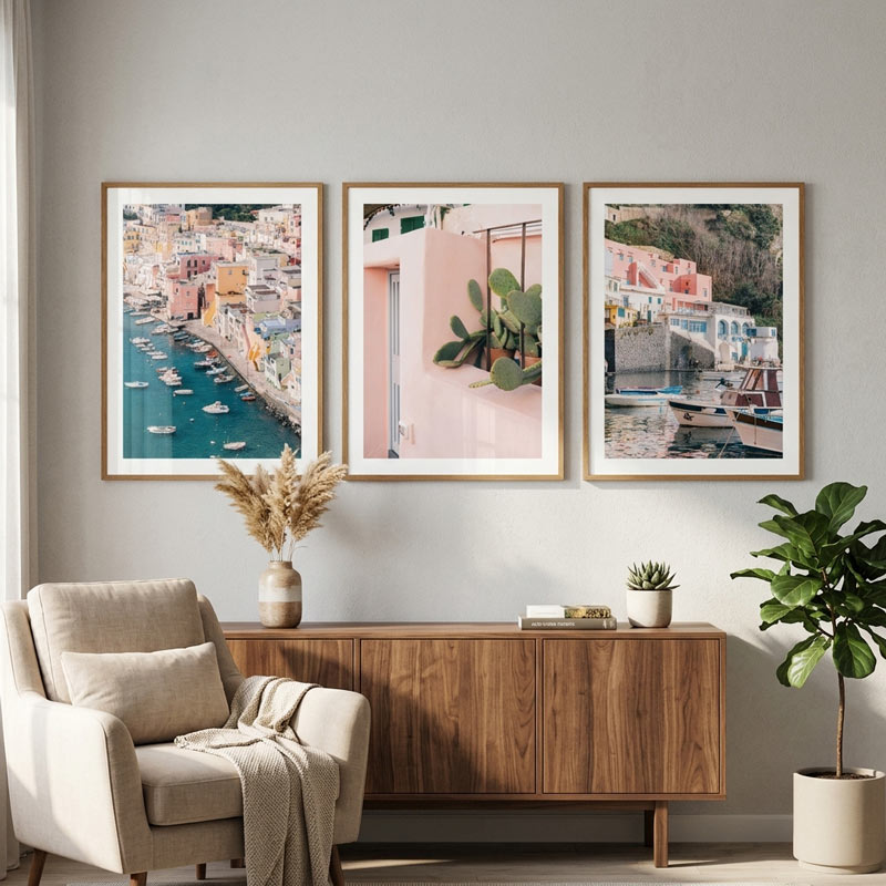Three framed art prints depicting coastal cityscapes and architecture are displayed on a light gray wall above a wooden sideboard in a styled living room with a beige armchair, pampas grass, and a potted fiddle-leaf fig.