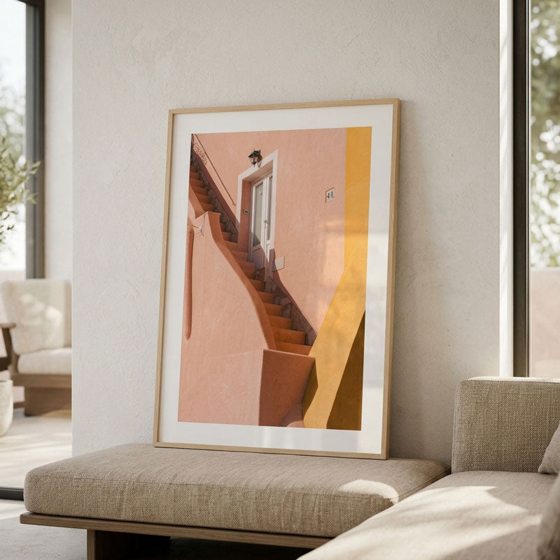 A framed art print featuring a minimalist architectural scene of a staircase and door on a peach and yellow background, leaning against a wall in a modern living room setting.