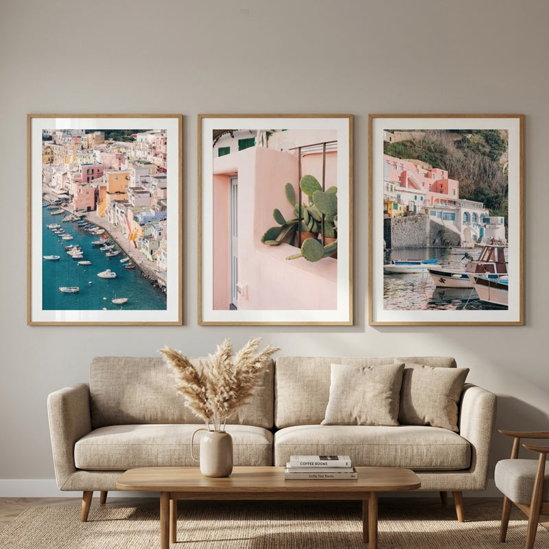 Three framed art prints depicting coastal village scenes and a pink wall with a cactus, displayed in a light wood frame on a beige wall above a modern beige sofa in a styled living room.